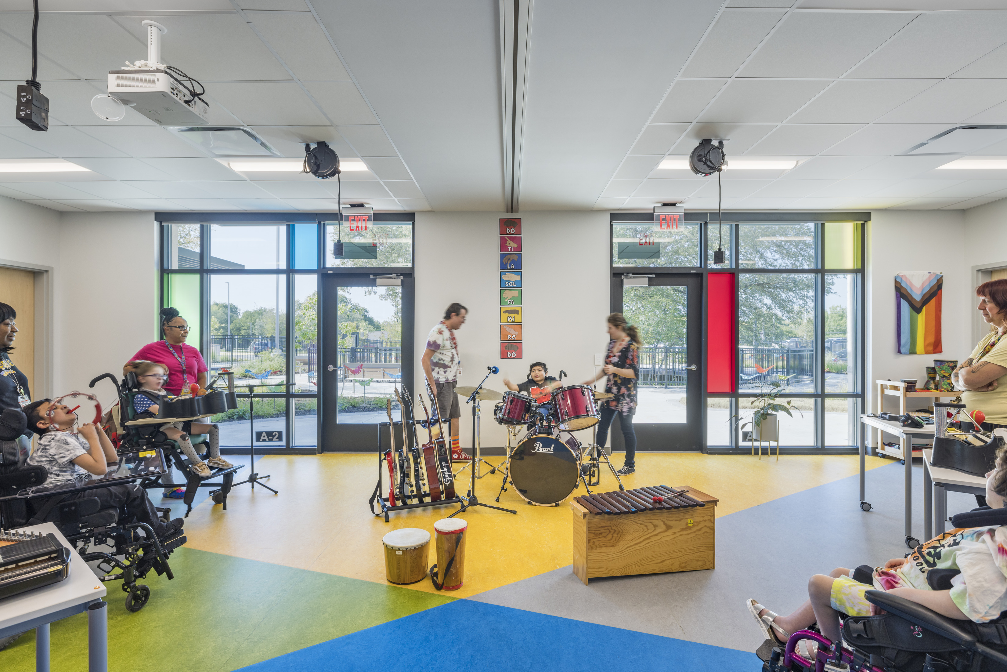  A brightly lit, accessible music room where children with diverse abilities are engaged in playing musical instruments. The room features vibrant, multicolored flooring, large windows with colored panels, and various musical instruments like drums, guitars, and tambourines. Teachers and staff assist the children, creating an inclusive and interactive learning environment.