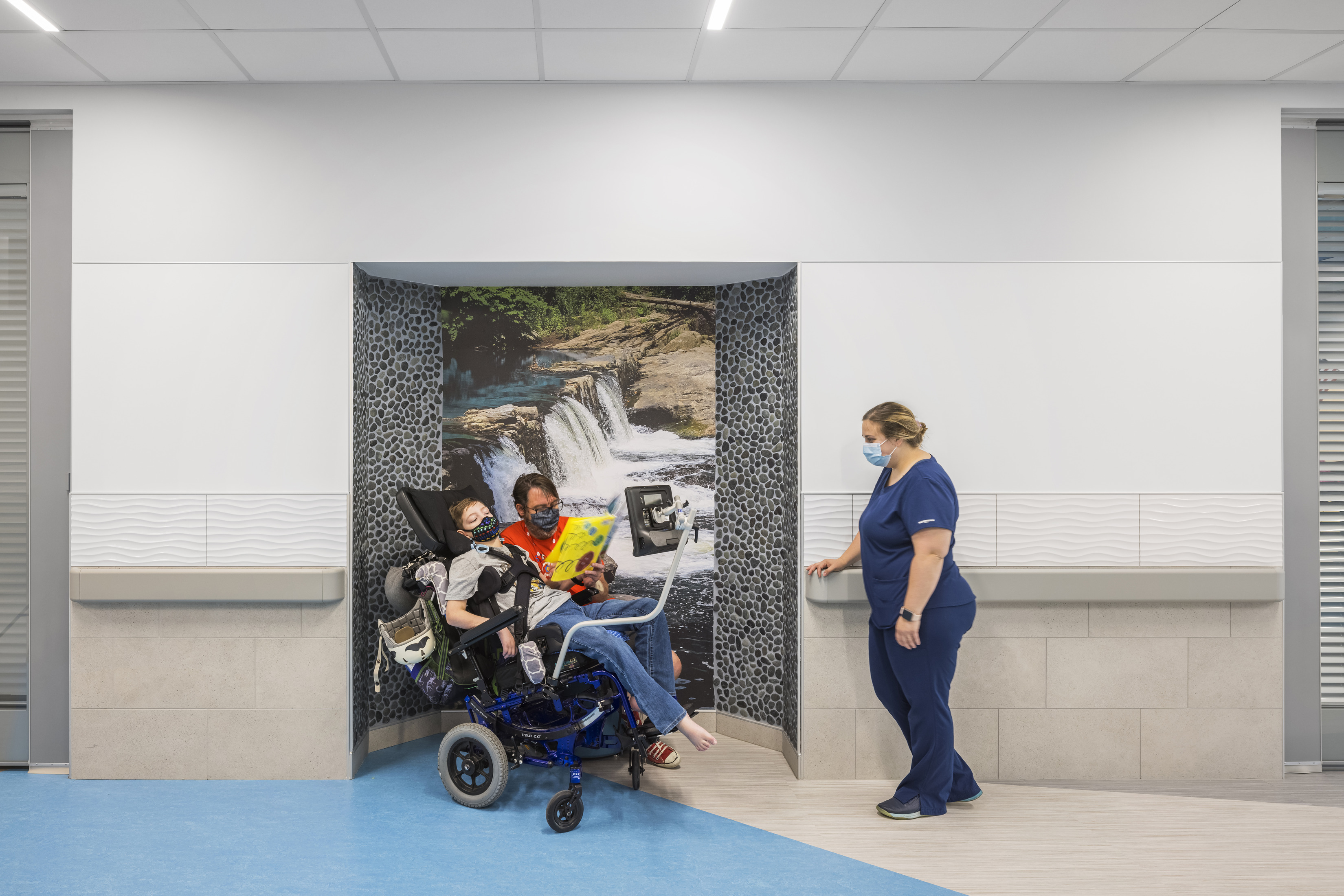 A nurse interacts with a child in a wheelchair in front of a wall mural depicting a peaceful waterfall scene. 