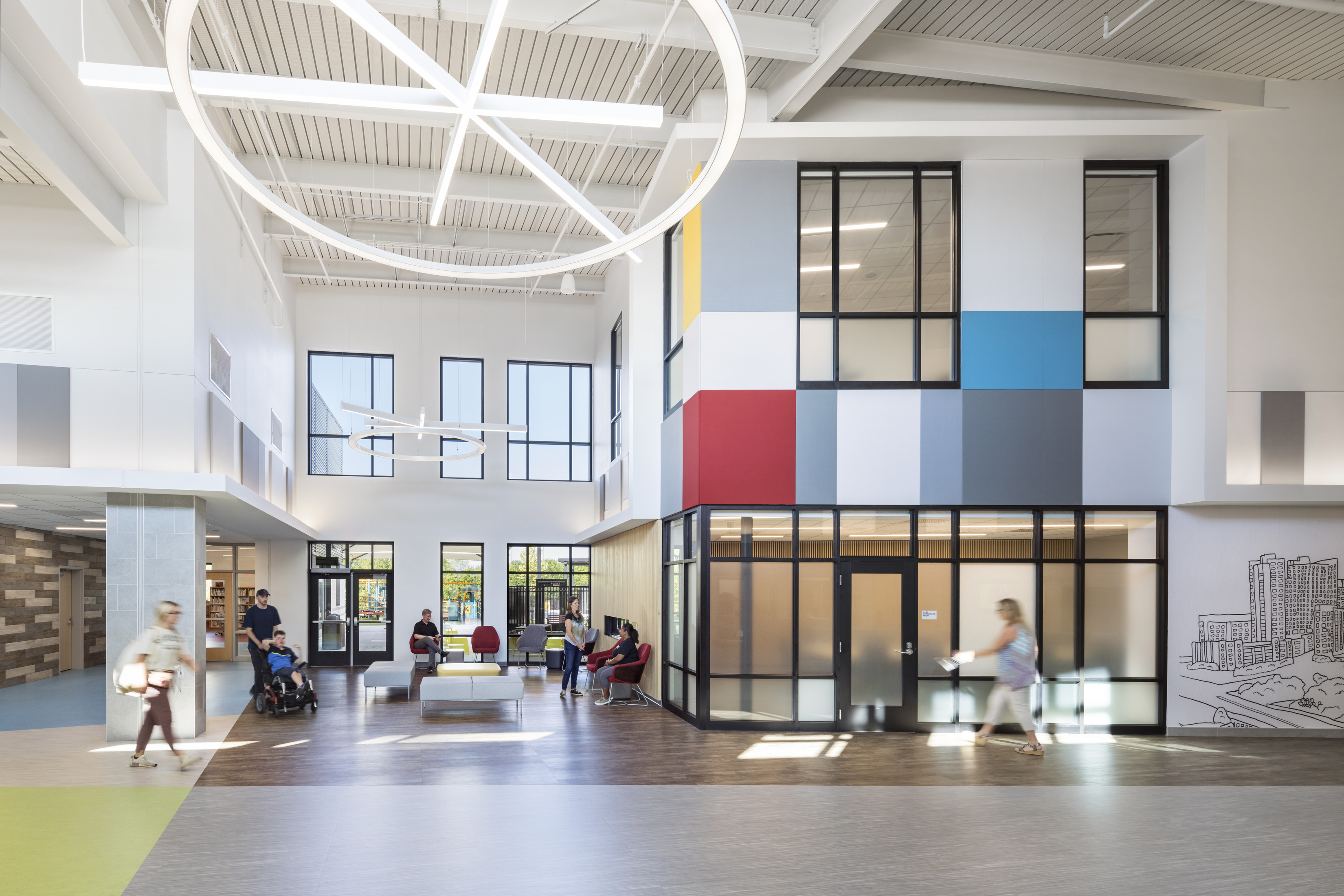  A spacious, open atrium with high ceilings and large windows that allow natural light to fill the space. The room has a modern design with a colorful panel wall, lounge seating areas, and people interacting and moving around. The flooring is patterned in various textures and colors, creating a dynamic yet welcoming atmosphere.