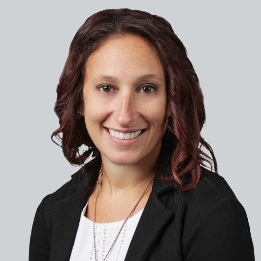 Nora Colman Headshot - short, curly, brown hair, brown eyes, wearing a black blazer and white shirt