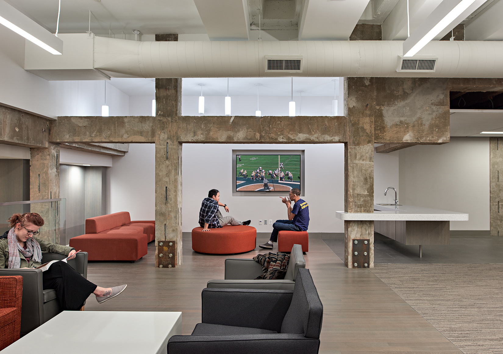 A modern, industrial-style lounge area within a larger building. The space is characterized by exposed concrete beams and columns, which add a raw, unfinished look to the otherwise sleek environment. In the center, two individuals are seated on orange circular sofas, engaged in conversation with a large television screen mounted on the wall behind them. The room is furnished with comfortable chairs and sofas in neutral tones of gray and orange. Above, exposed ductwork and suspended light fixtures contribute to the industrial aesthetic. To the right, there's a small kitchenette area with a sink, suggesting this space is designed for informal gatherings, discussions, or relaxation.