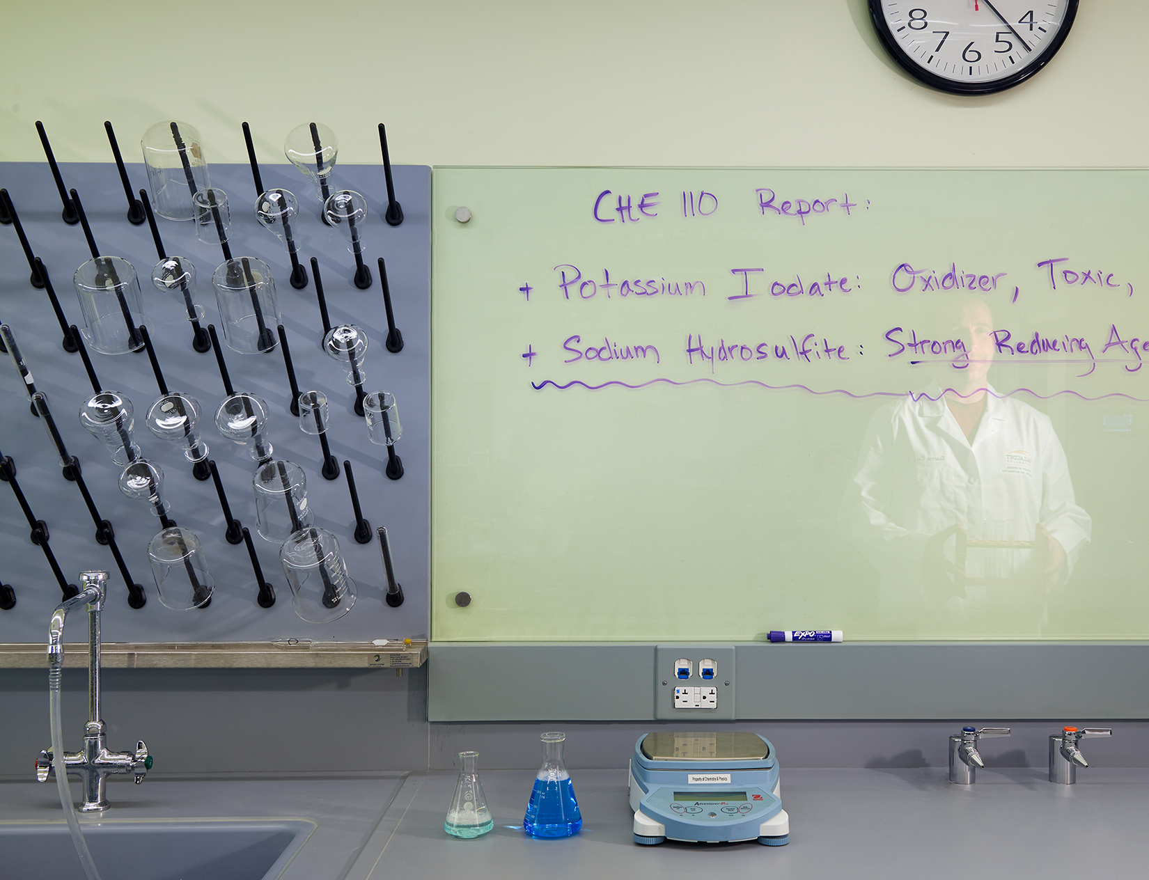 The image showcases a clean, organized science laboratory environment. On the left, a drying rack holds an assortment of glassware, including beakers and flasks, all arranged neatly. A countertop below the rack is equipped with a sink, a piece of lab equipment, and a couple of conical flasks filled with colored liquids. On the right side, a large glass whiteboard displays handwritten notes about a chemistry report, detailing the properties of potassium iodate and sodium hydrosulfite. The reflection of a person in a lab coat is faintly visible on the whiteboard.